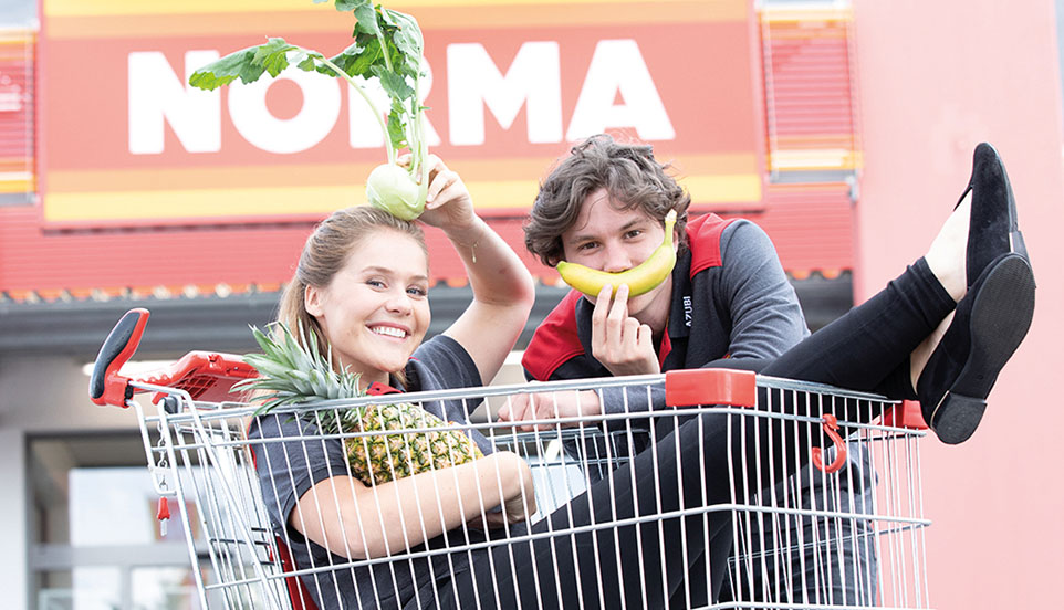 Mädchen sitzt in einen Einkaufskorb mit Obst und Gemüse, im Hintergrund NORMA-Filiale