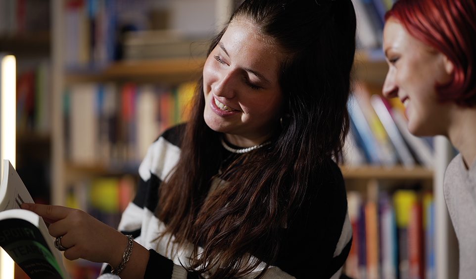 Zwei junge Frauen schauen vor Bücherwand gemeinsam in ein Buch