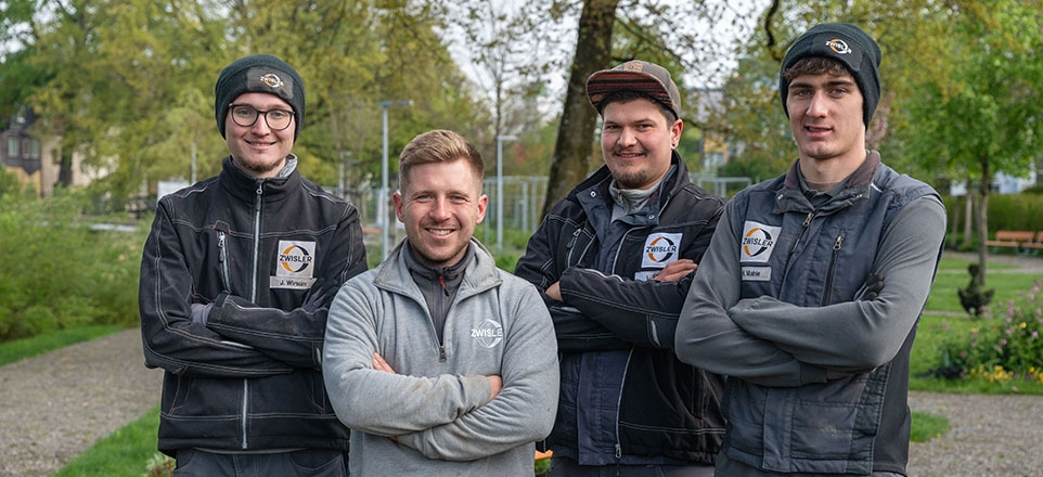 Vier junge Männer vor einer grünen Gartenanlage.