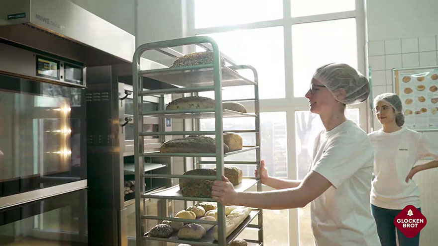 Junge Bäckerei-Mitarbeiterin mit Backwaren vor einem Backofen.