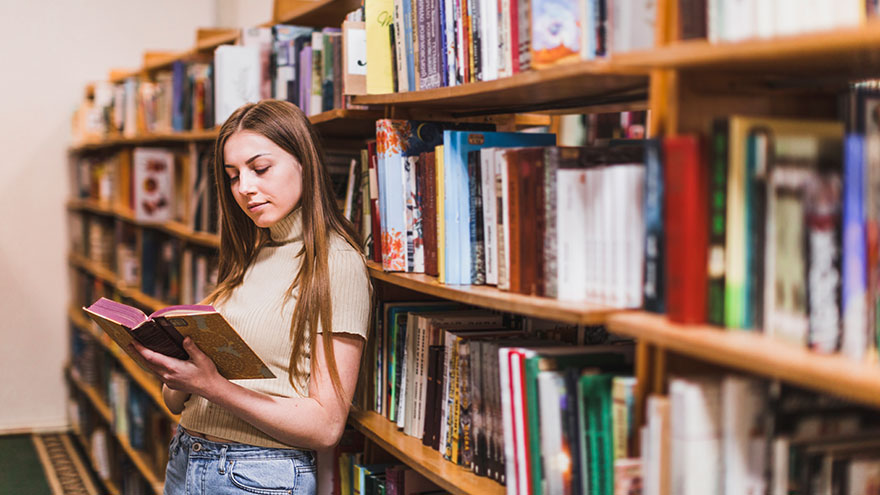 Junge Frau liest vor einem Bücherregal stehend. Bild von Freepik.